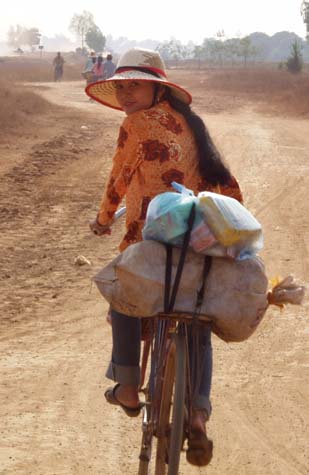 A Khmer lady is cycling along the side of a dusty road. The road itself is in such poor condition that the local cyclists have created a second route paralleling the first. Her bike rack is loaded with food and a white sack with unknown contents. The bare brown earth of the path and the surrounding area stretch off towards distant mountains, passing a few scattered trees on their way