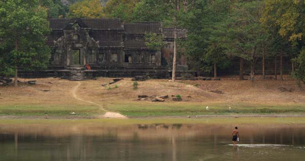 Photograph of a man carrying water across Angkor Wat's main moat in the dry season to a monk sitting on the steps of an entrance point to the main grounds