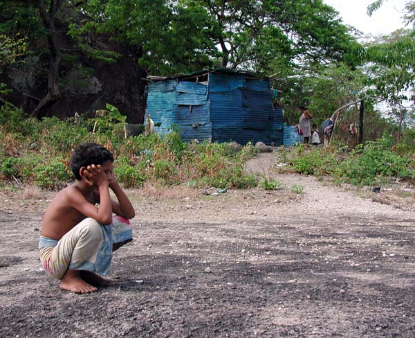 Photograph of a child crouched, head in hands, in front of the shack he lives in with his 8 siblings. The shack is roughly made from corrugated metal, his pregnant mother is standing next to the shack in the background