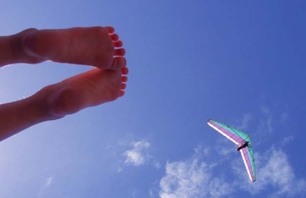 A pair of children's feet are in the foreground of the left hand side of the photo set against a blue sky. A hang glider is in the bottom left of the image passing under some whisps of cloud.