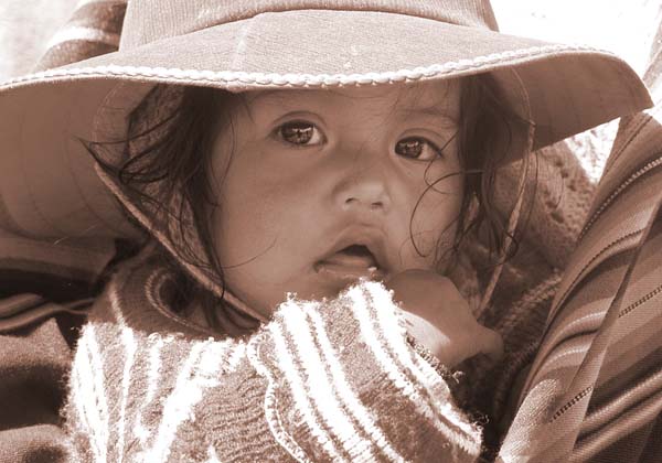 Photograph of a young child in a traditional, woven peruvian back carrier, she looks directly into the lense