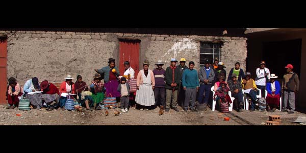 Photograph of the people at the community center in Palca. To the far left a group of three women are hiding their faces from the camera. In the center a lady stands proudly for the camera in her bright pink cardigan and brown bowler hat. To the right of her as we look at the image the men of the town are lined up in their working clothes.