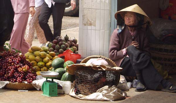 Photograph on an old vietnamese lady sitting against a wall in a traditional hat. People are pasing without looking at the fruit she is trying to sell