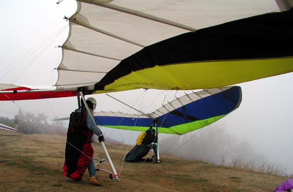 Two hang gliders wait on a hill for a chance to launch while a heavy fog obscures the trees in the background of the shot. There are glimpses of others waiting their turn.
