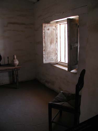 Photographic still life of a room with a wooden shuttered window open allowing light to enter the gloom. A vase and candlestick sit on a table against the facing wall, a chair against the wall containing the window that runs towards the camera on the right of the image