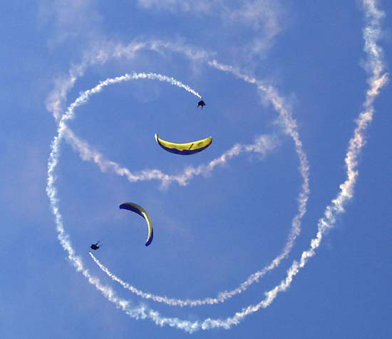 Two paragliders spiral down towards earth on opposite sides of an invisible circle, smoke streaming behind them.