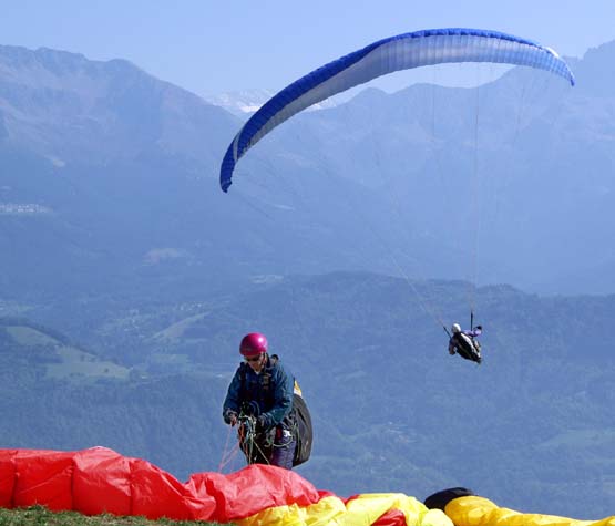 Photograph of a paraglider preparing to launch his red wing, a bright yellow wing rests on the ground to the right of the image. Already airborne a blue wing is set against the dramatic St Hilaire mountains and woodland.