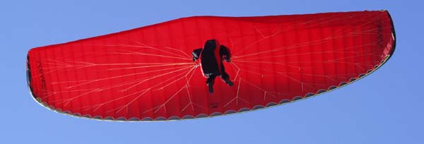 A paraglider on a bright red wing passes directly overhead, contrasted against a perfectly blue sky