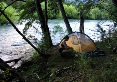 Dana squats in front of the tent working. The tent is along the banks of the Rio Manso, the water in the background is fast flowing. 3 tree trunks are just behind the tent, it is evening twilight.